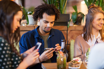 leisure, people and technology concept - happy friends with smartphones eating and drinking at restaurant