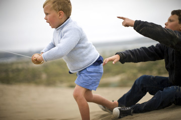 Young boy looks up anxiously as he uses a single-string wooden kite reel  while his older brother sits behind him and points at the sky.
