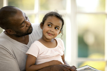 Young girl sitting on her father's lap while reading a book.