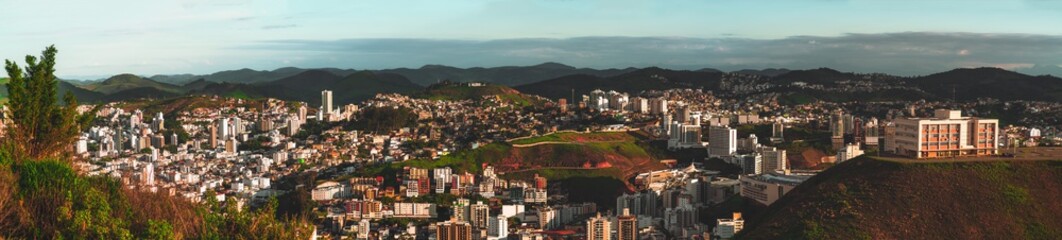 Fototapeta premium Wide panoramic view from a high point an urban landscape lit by bright evening sun: plenty of residential, office, educational houses, hills with favelas, mountain ridge in the distance; Juiz de Fora