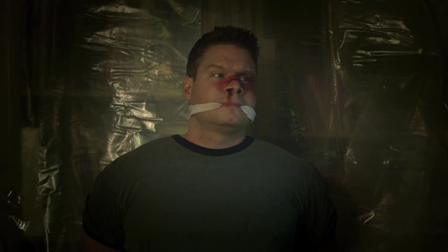 A tied-up man that is beaten and bruised, sees something thats shocks him