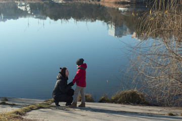 Fototapeta na wymiar On the shore of lake mom talks to her son. Family walking in the park near lake. Mother and son look at each other in spring. buildings are reflected in water