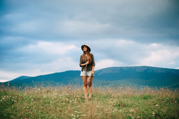 Naklejka premium Young girl in beige hat and casual clothes with long hair is walking with cloudy sky and green mountains on background