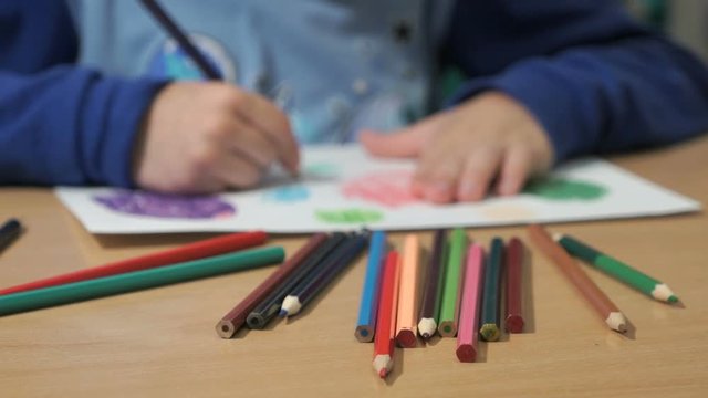 Close-up of hands of unidentified little boy in blue sweater sitting at table drawing pictures using multicolored pencils in kindergarten