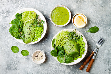 Obraz premium Vegan, detox Buddha bowl with avocado, spinach, micro greens, edamame beans, zucchini noodles and herb green dressing. Top view, grey concrete background