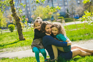 Little girl hugs mother sitting on the grass. Child, playing, threw his mother on grass. Family in spring walks on street.