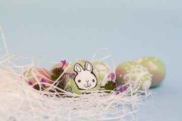 Easter background in pastel colors: handmade hand painted eggs in pink and green gradient, decorative bunny doodles, white straw, blue backdrop. Bright colorful composition, blurred, selective focus.