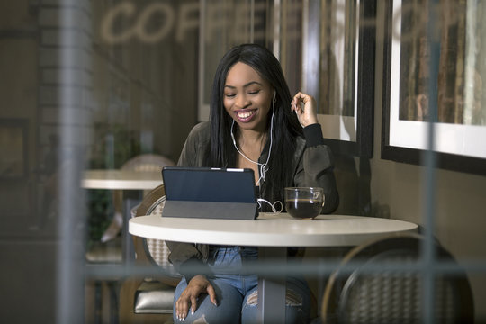 Window view of a black female wearing headphones and watching videos on a tablet in a coffeeshop streaming via 4g or 5g wifi internet.  She is sitting and holding a cup of coffee