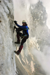 Climber with an ice ax climbs on a vertical ice wall. He is wearing equipment. Helmet, rope, crampons and ice ax. On the background there are mountain ridge, a lot of ice, clouds and rocks.