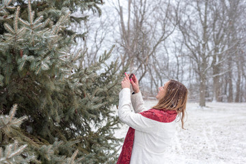 happy woman taking picture of snow falling on pine tree with smart phone