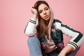 Portrait of a beautiful model in a sports jacket print and jeans sitting on a pink background