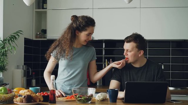 Cheerful couple at table woman dancing and cooking while gives hasband to try red pepper, man sitting working on laptop, he shows something funny on screen