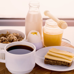 healthy breakfast fresh cereal, cheese sandwich, hard boiled egg, milk bottle, coffee cup and honey jar, toned warm sunny morning light