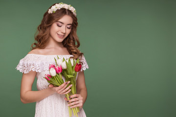 A beautiful young woman stands on a purple background, wearing a white dress and a wreath of flowers on her head, and a bouquet of tulips. International Women's Day, March 8.