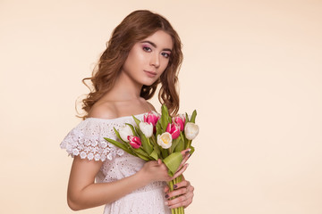 Beautiful young woman stands on a beige background, in a white dress and a bouquet of tulips. International Women's Day, March 8. Copy-space.