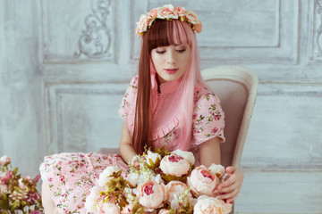 Beautiful young woman doll in a pink dress in a room with flowers, sitting on a chair by the...