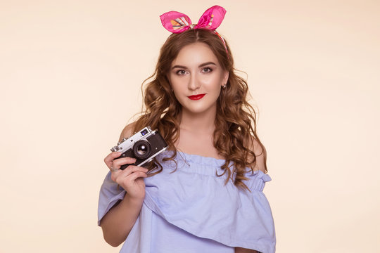 A beautiful young girl in a pin-up image takes pictures on an old DSLR, on a beige background. Metal camera.