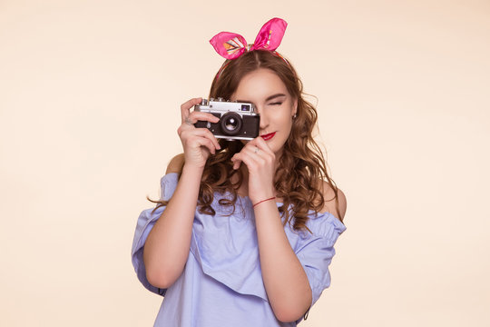 A beautiful young girl in a pin-up image takes pictures on an old DSLR, on a beige background. Metal camera.