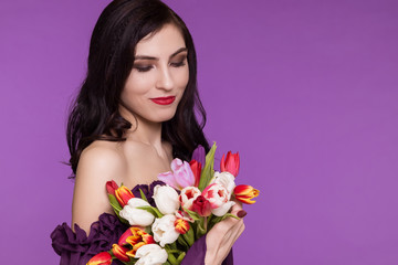Beautiful young woman, brunette, stands on a purple background, in an air dress and a bouquet of tulips. International Women's Day, March 8. Copy space.