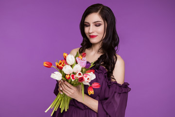 Beautiful young woman, brunette, stands on a purple background, in an air dress and a bouquet of tulips. International Women's Day, March 8.