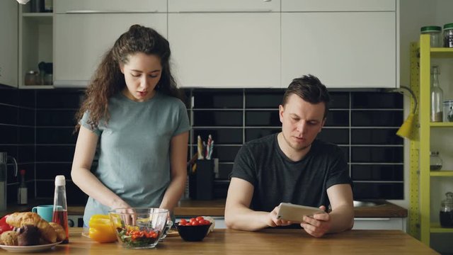 Attractive couple in the kitchen. Man playing video game on smartphone while his girlfriend cooking