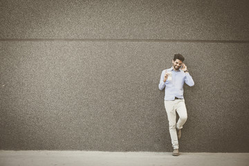 Young businessman leaning against wall with smart phone. Man holding cup of coffee.