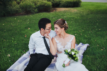 The bride and groom are smiling, sitting on the grass and plaid. International wedding. Asian and...