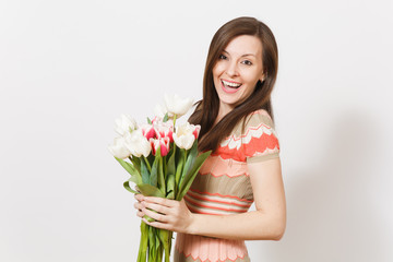 Beautiful young happy brunette female in light patterned dress is holding bouquet of white and pink tulips in hands, smiling and rejoices isolated on white background. Concept of holiday, good mood.