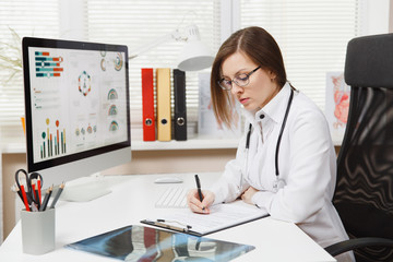 Young woman sitting at desk, working on computer, filling out medical documents in light office in hospital. Female doctor in medical gown, stethoscope in consulting room. Healthcare, medicine concept
