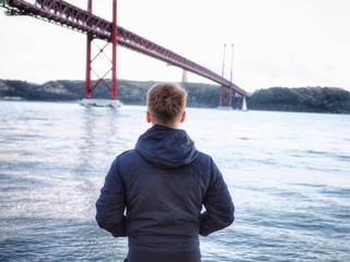 A man in a jacket stands against the background of the sea city and the bridge in  Lisbon.