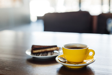 A horizontal image of a yellow cup of black coffee and a plate with a piece of chocolate cake. Bokeh background.