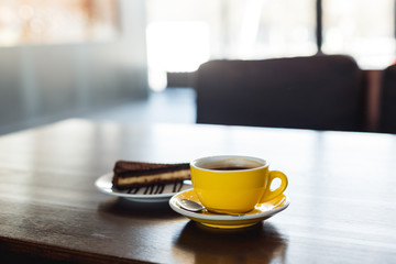 A horizontal image of a yellow cup of black coffee and a plate with a piece of chocolate cake. Bokeh background.