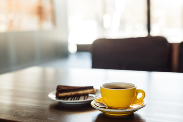 A horizontal image of a yellow cup of black coffee and a plate with a piece of chocolate cake. Bokeh background.