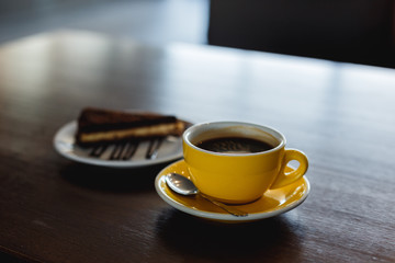 A horizontal image of a yellow cup of black coffee and a plate with a piece of chocolate cake. Bokeh background.
