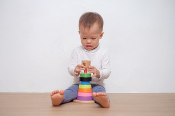 Cute little Asian 18 months / 1 year old toddler baby boy child play with colorful wooden pyramid toy / stacking ring toy. Kid playing with educational toy isolated on white wall, with copy space
