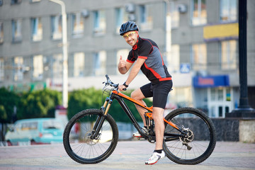 Fototapeta premium Sporty smiling guy biker in cycling clothes and helmet on bicycle, looking to the camera, showing thumbs up. Man resting and relaxing after work, training on bicycle on city streets. Healthy lifestyle