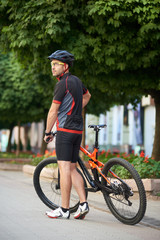 Back view of sporty bicyclist posing near bicycle shooting for sport ad cycling garment campaign. Young male admiring city while taking break riding bike on morning race.