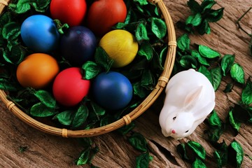Happy Easter Day! White rabbit and colors Easter eggs in basket with green flower decoration on wooden table background top view