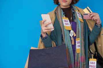 Asian shopaholic woman in clothes with sale price tags using application on her smartphone - obrazy, fototapety, plakaty