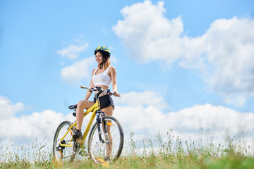 Obraz premium Happy young female rider cycling on yellow bicycle on a grass, wearing helmet, enjoying sunny day in the mountains against blue sky with clouds. Outdoor sport activity, lifestyle concept. Copy space