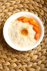 Low-calorie porridge for a healthy diet and lifestyle. Barley with honey and bits of orange pumpkin in a deep white plate. Dark background, wooden table and wicker stand