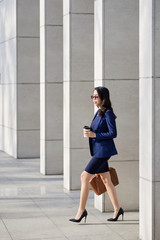Profile view of confident young manager in formalwear holding paper cup of fragrant coffee in hand while leaving office building after hard working day