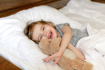 Active adorable baby wakes up lying in bed with her Teddy bear toy, soft white linens. Children's...