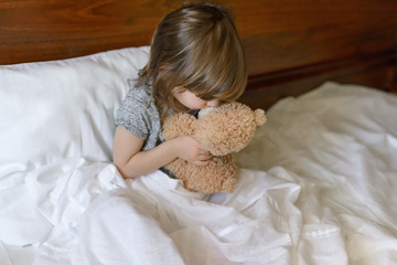 Active adorable baby wakes up lying in bed with her Teddy bear toy, soft white linens. Children's day. World Smile Day.