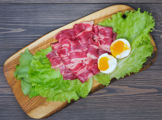 Raw bacon from marbled beef meat on lettuce sheets on an olive wood board on a dark background of natural wood with an egg in a crumpled