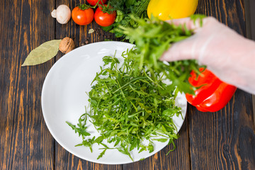 High angle view of vegetables, unrecognizable hand in glove puts arugula on white plate