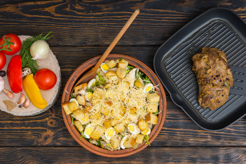 High angle view of large clay dish with tasty caesar salad, prepared meat on a grill pan next to vegetables on wooden desk