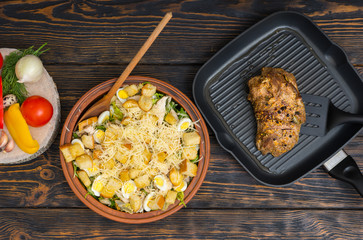 High angle view of large clay dish with tasty caesar salad, prepared meat on a grill pan with kitchen spatula next to vegetables on wooden desk