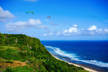 Paragliding over the ocean on the island of Bali. The blue sky shimmers with the blue ocean. The parasutists fly high above the precipice like birds. Aerial view with copy space