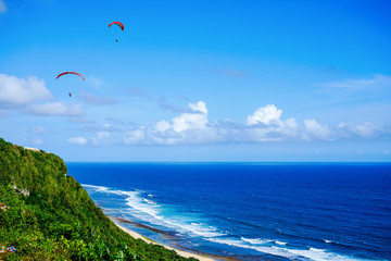 Paragliding over the ocean on the island of Bali. The blue sky shimmers with the blue ocean. The parasutists fly high above the precipice like birds. Aerial view with copy space